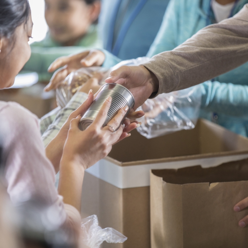 Young woman receives a canned food item from food bank. An unrecognizable volunteer hands her the canned food item.